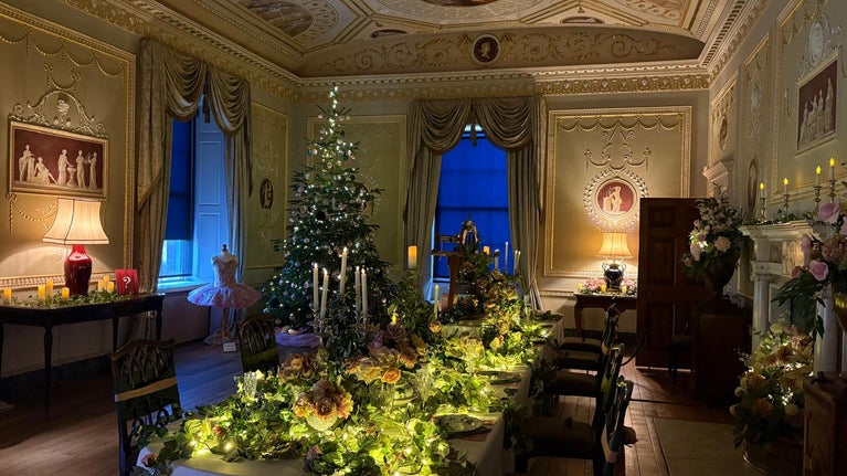 Dining room decorated for Christmas with a dining table covered in ivy, pink flowers and candlesticks. In the background is a Christmas tree decorated in green and pink baubles with ivy.